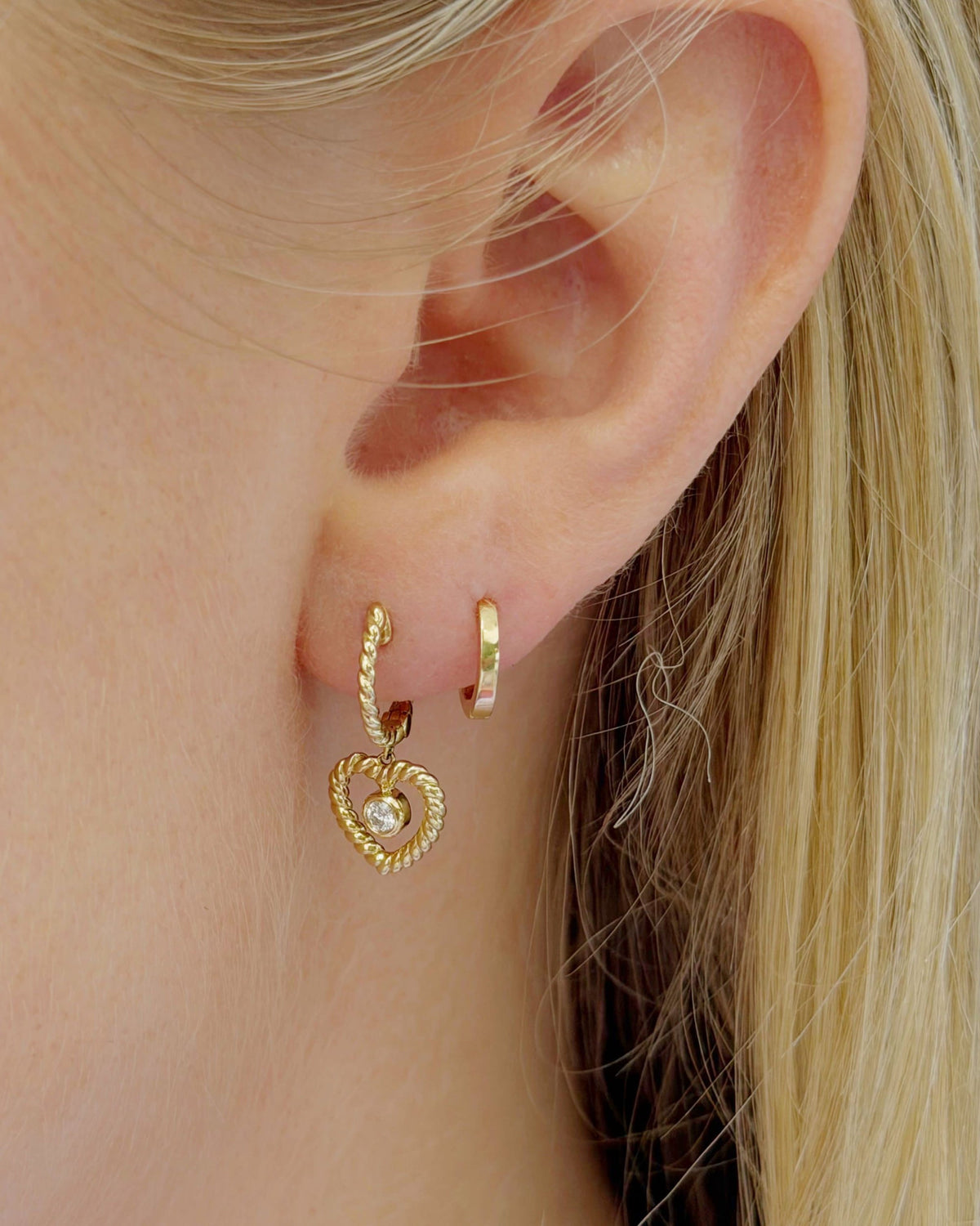 Petite Hoop in a close-up ear stack next to a twisted heart charm hoop, blonde hair behind.