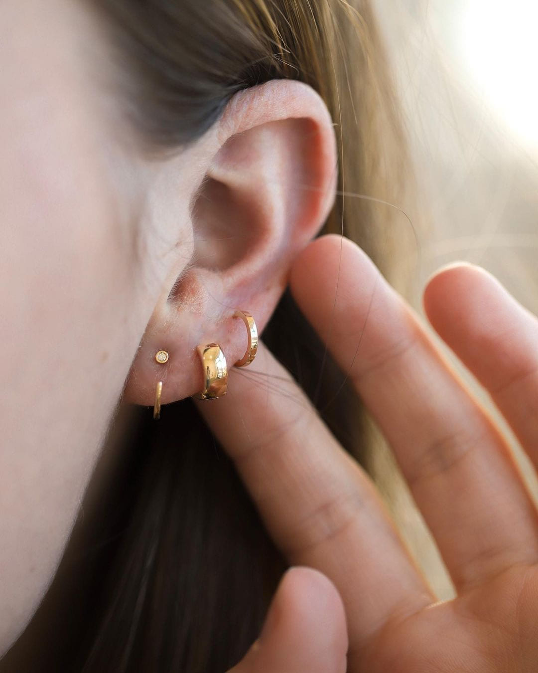 Petite Hoop in an ear stack with a chunky dome huggie and a tiny stud, fingertips behind the ear.
