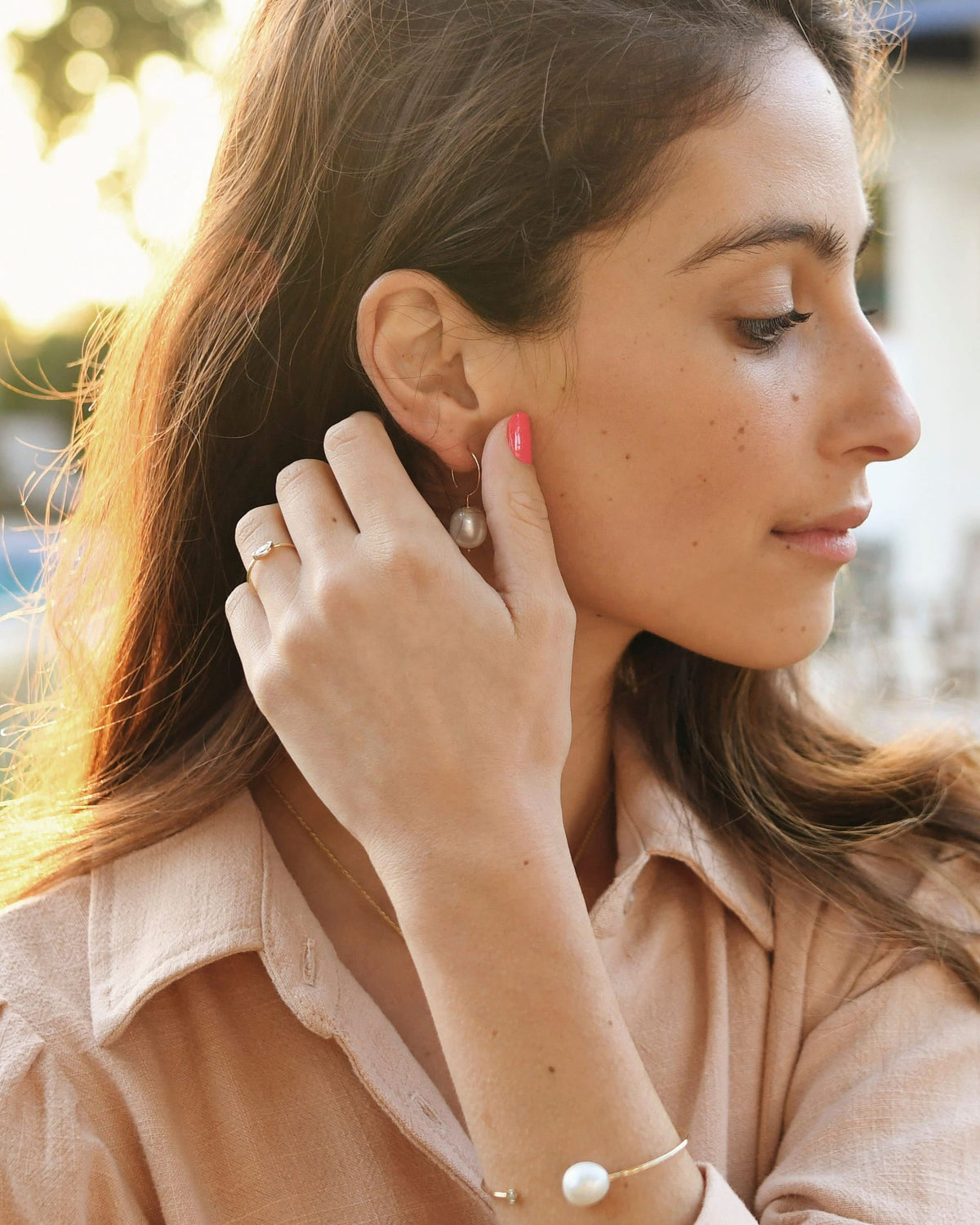 Baroque Pearl Swing Hoop on a model in a blush shirt, fingertips at her ear in warm sunset light.