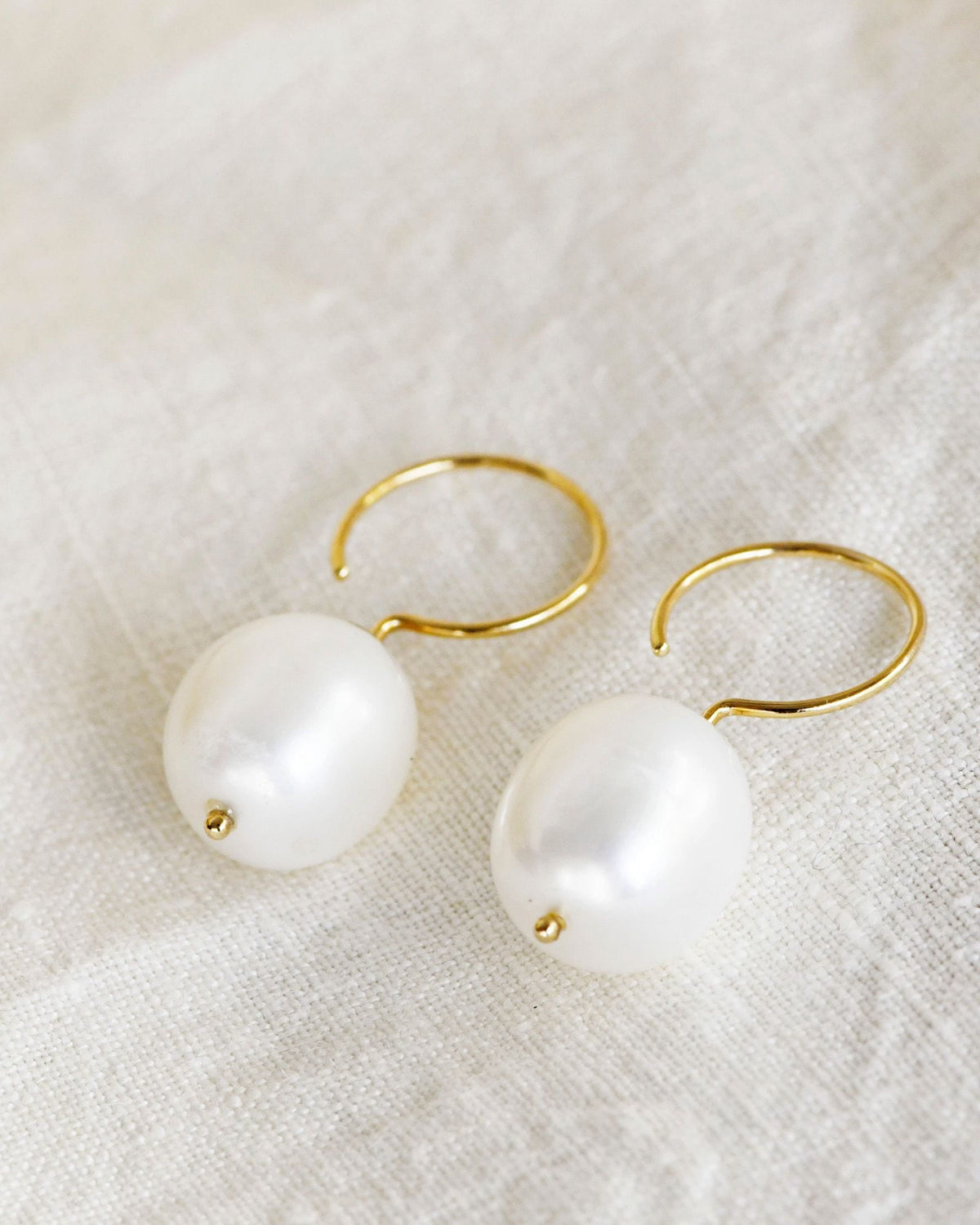 Baroque Pearl Swing Hoops, a pair resting on white linen with their open gold hoops showing.