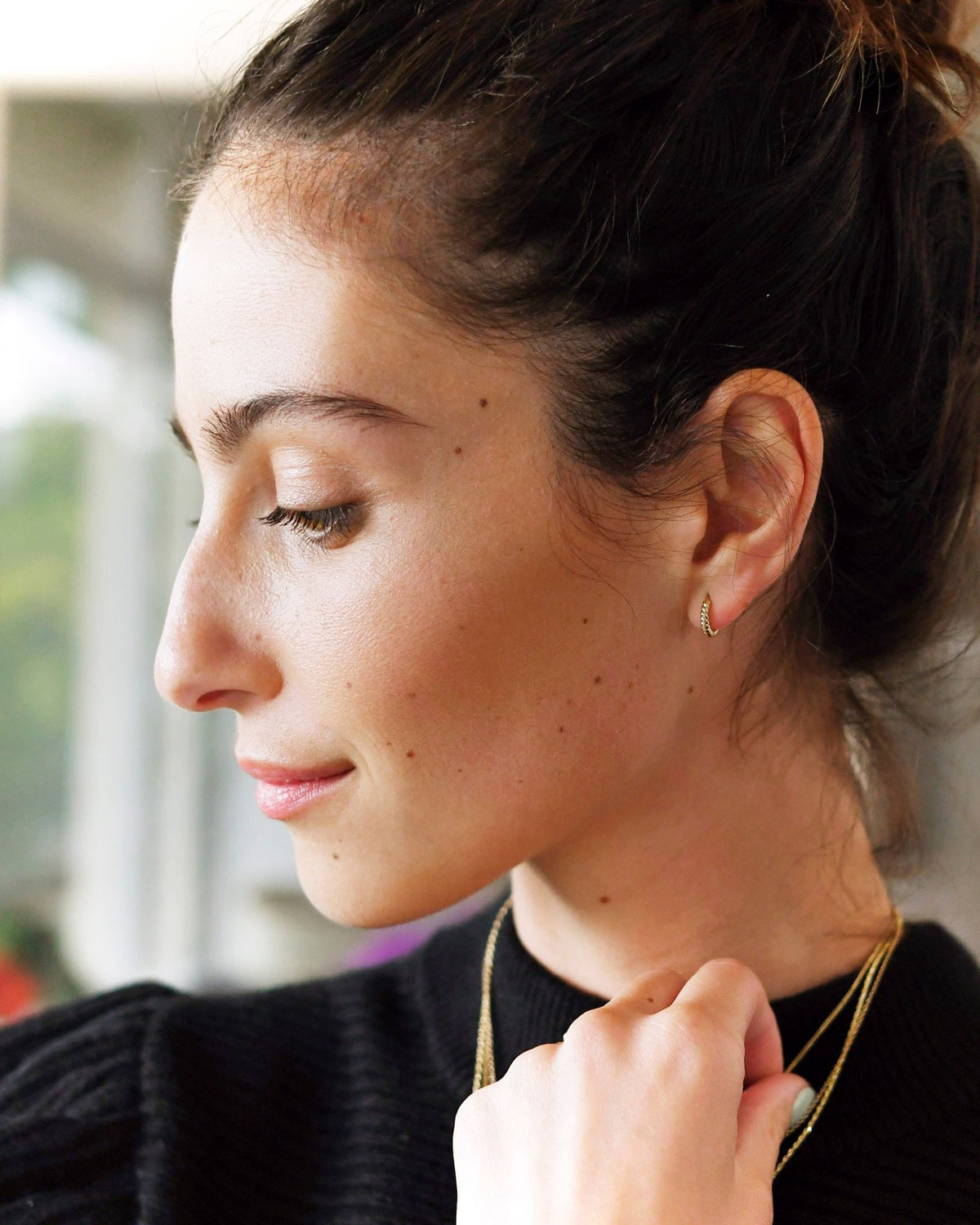 Twisted Petite Hoop on a model in profile with a messy bun and a black sweater, her hand at her necklace.