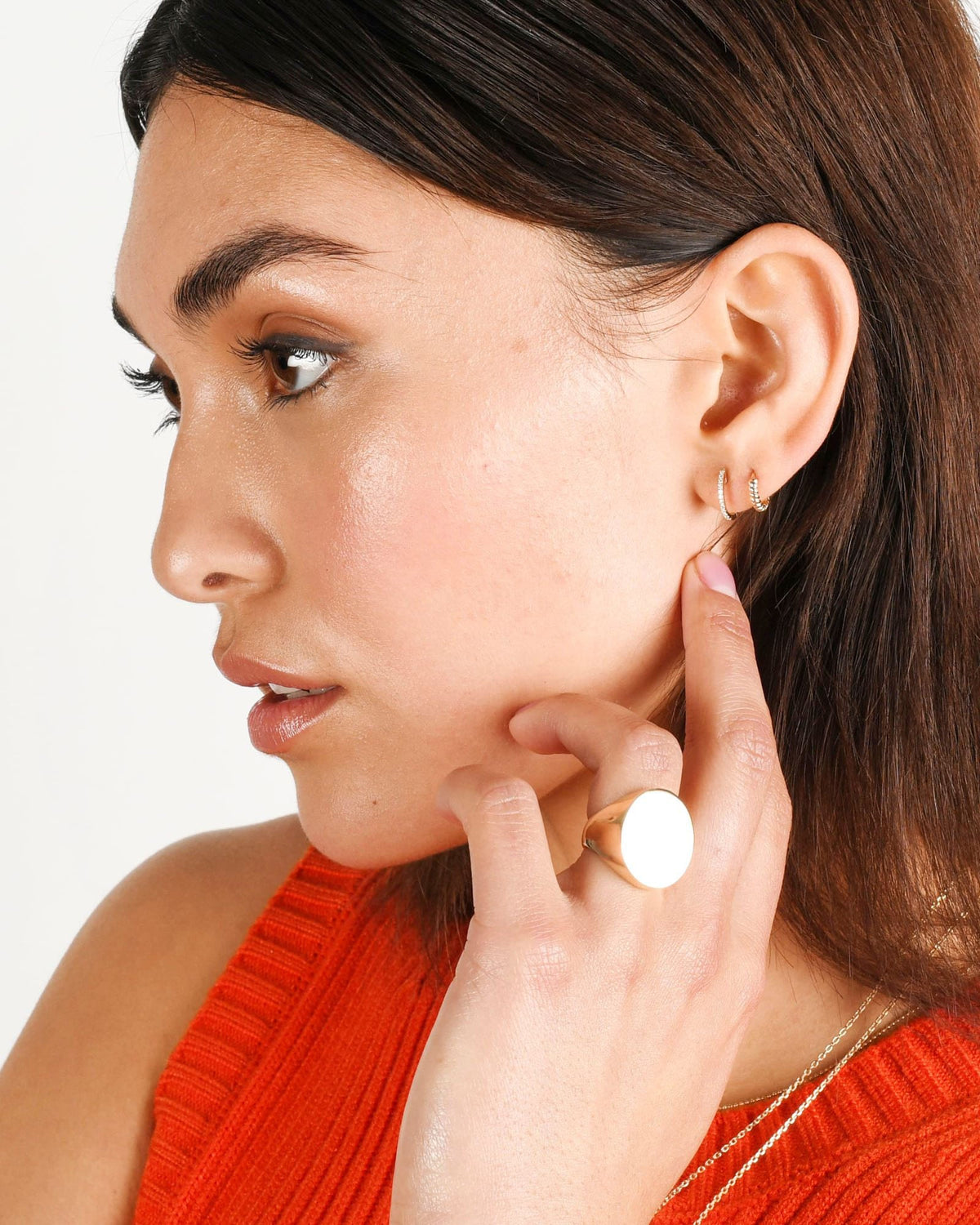 Twisted Petite Hoop beside a pave huggie on a model in an orange knit top, her finger at her jaw.