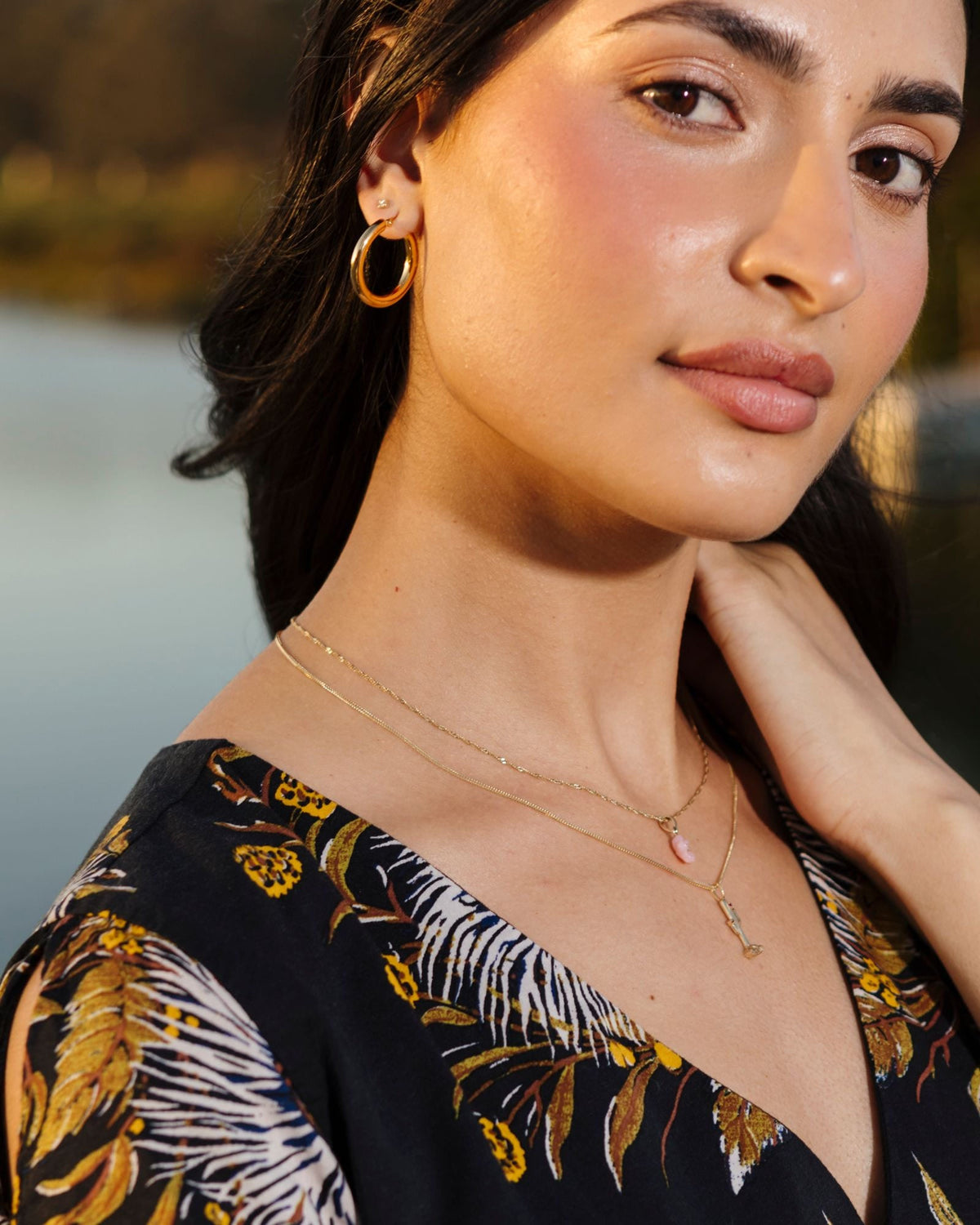 Junior Helium Hoop on a model in a black floral dress by the water, her hand at her neck.