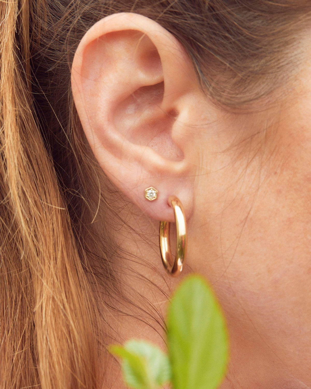 Diamond Hex Stud in close-up on the earlobe next to a chunky gold hoop, with green leaves in front.