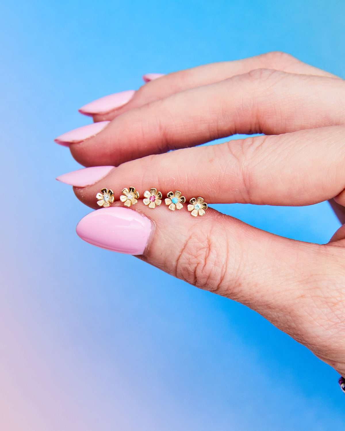 Fingertip holding In Bloom Birthstone Studs against a blue and pink gradient.