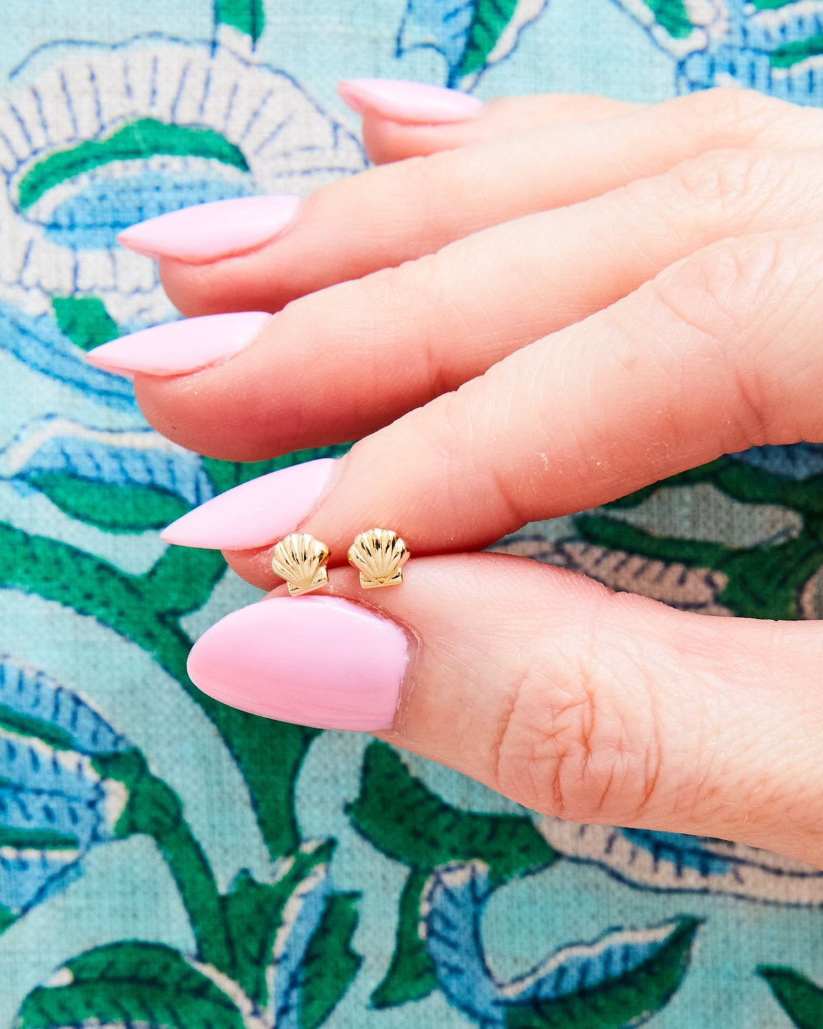 Fingertip holding Petite Scallop Shell Studs against a green and blue block print.