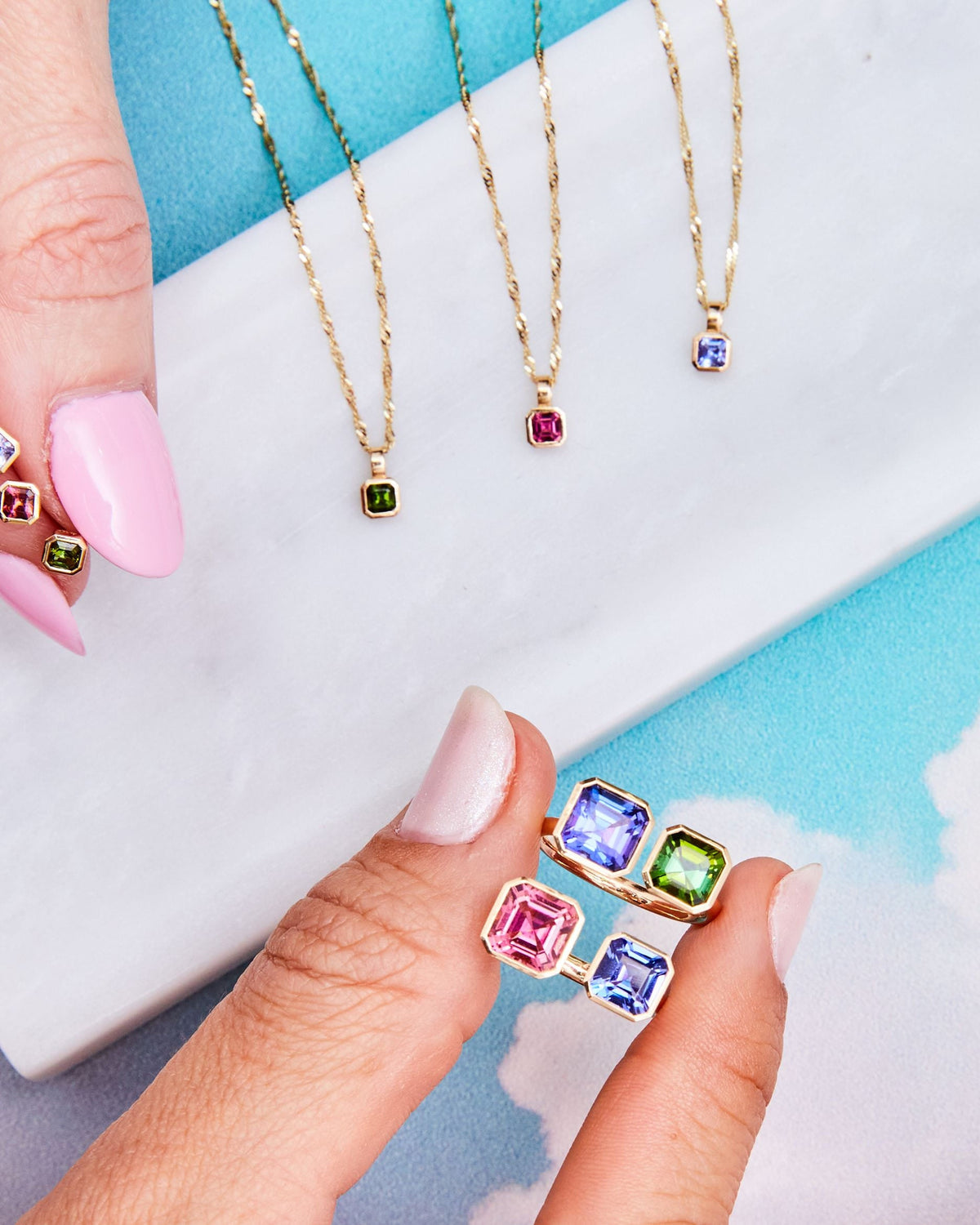 Fingertips holding Petite Asscher Studs in iris and other stones against clouds.