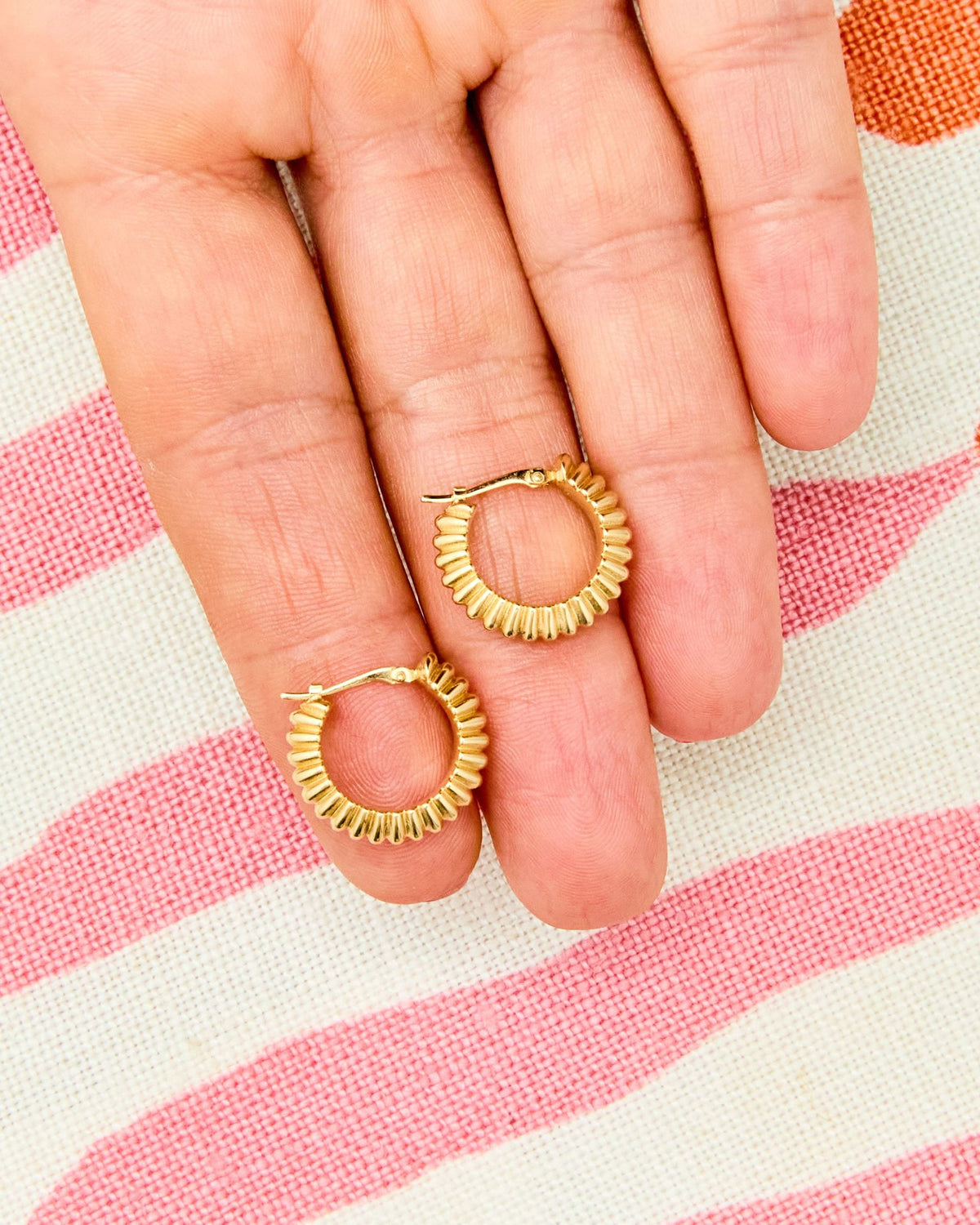 A pair of Ridged Knife Edge Helium Hoops on the fingers against pink stripes.