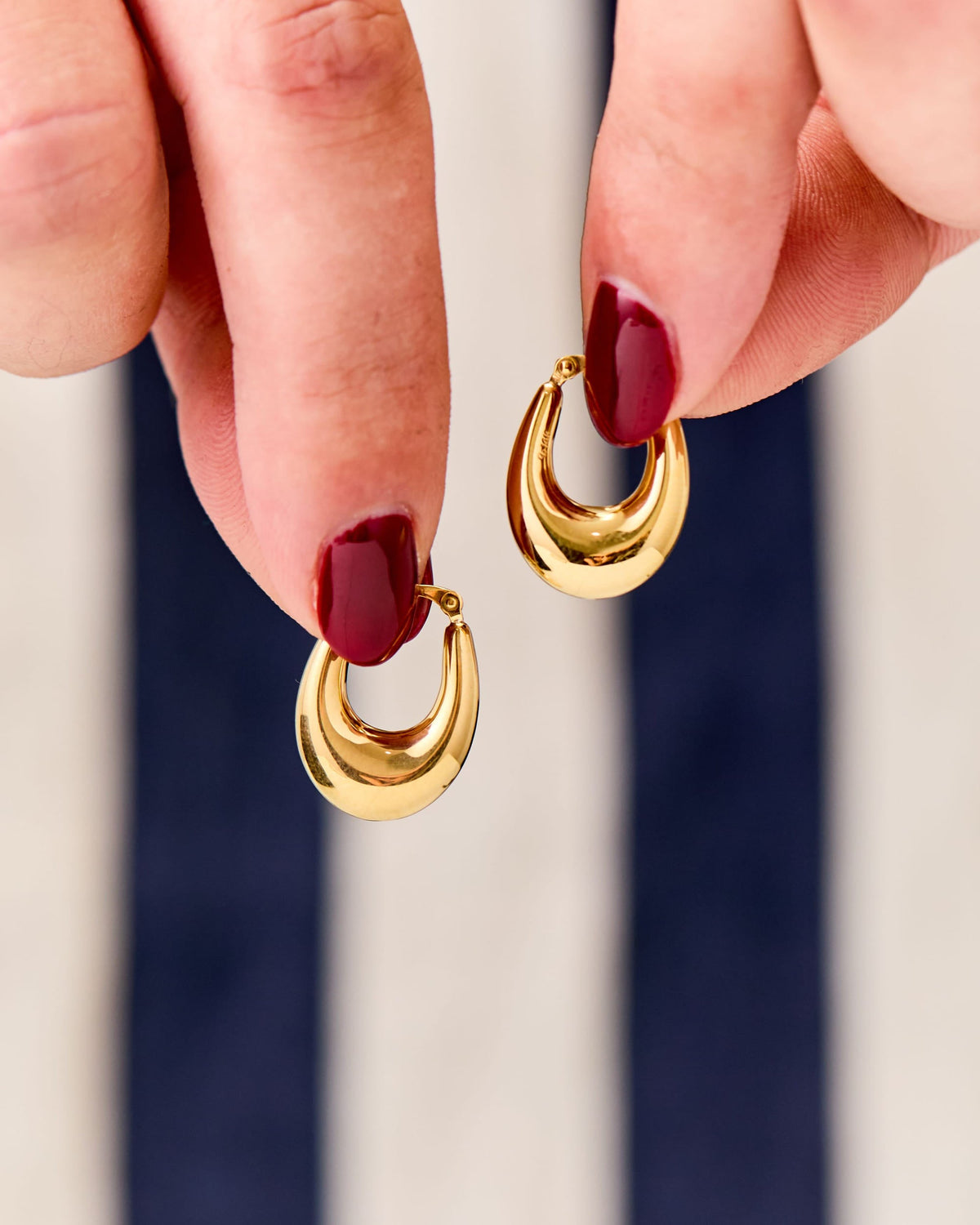 Fingers holding a pair of Junior Helium Teardrop Hoops against stripes.