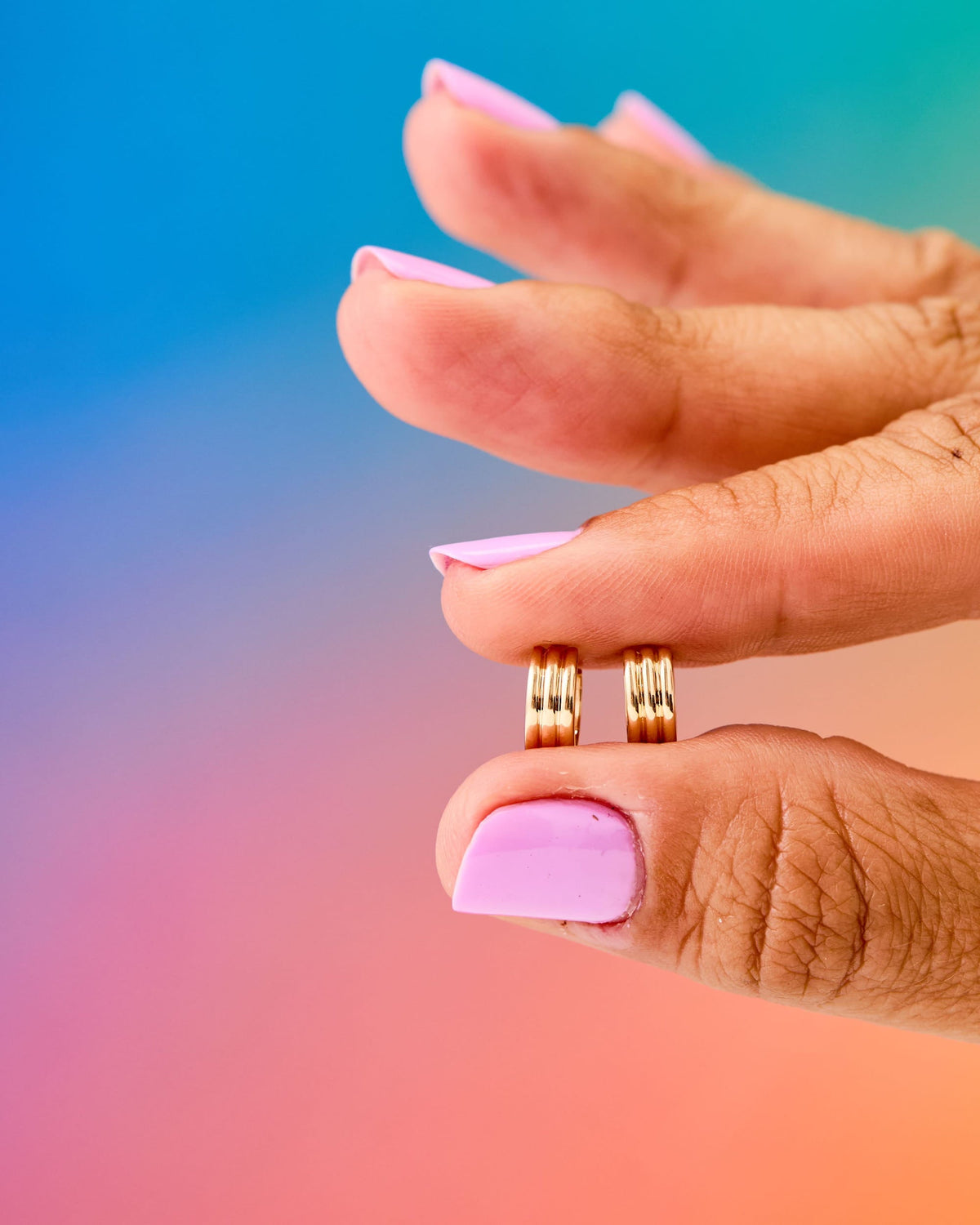 Fingers holding a pair of Mini Triptych Hoops against a rainbow gradient.