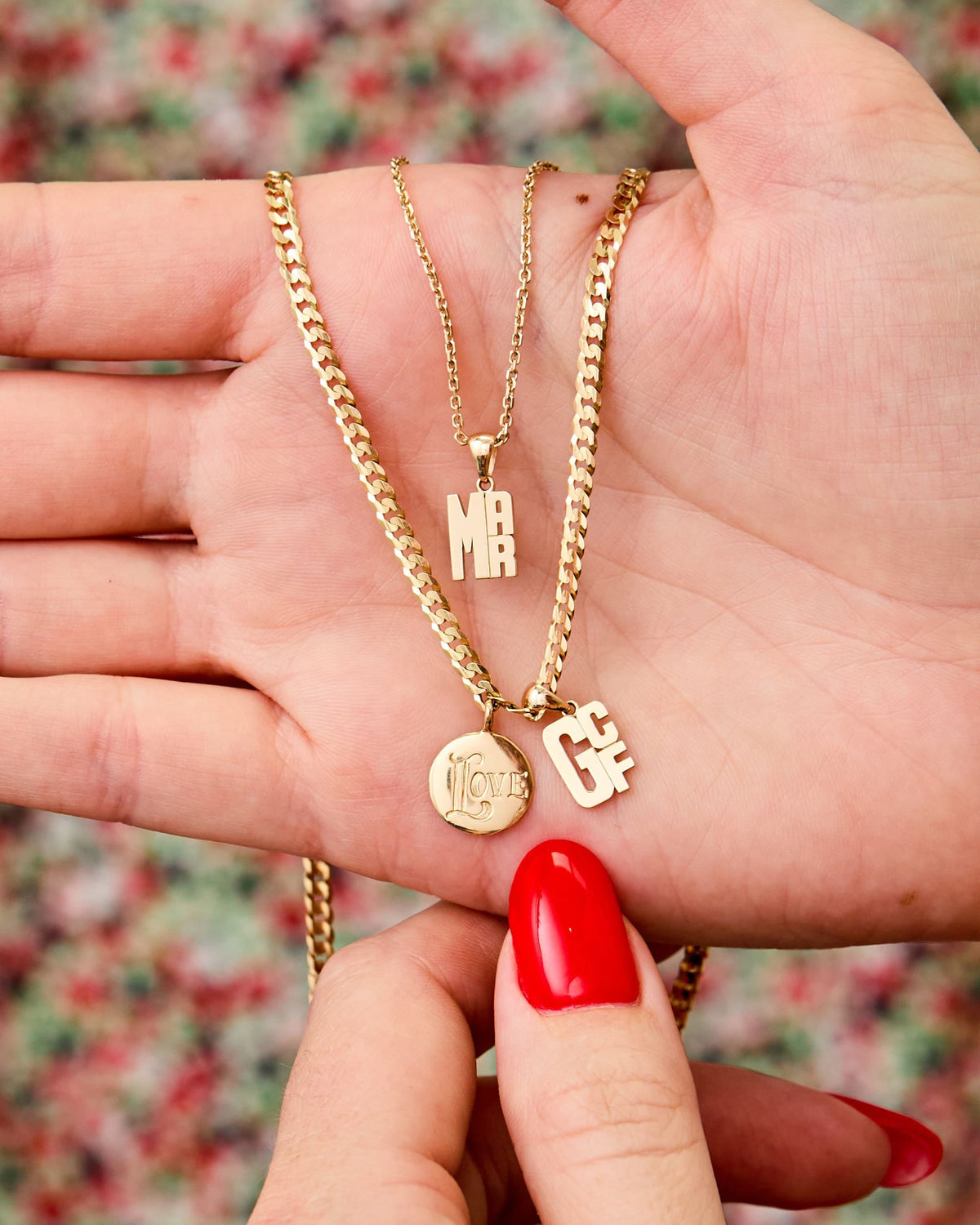 Hand holding gold Medallion Signet Necklaces and initial charms against a floral background.