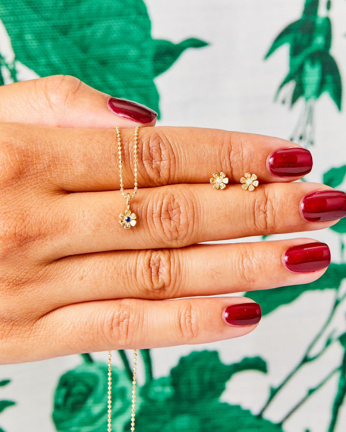 Hand with red nails showing an In Bloom Birthstone Charm and studs over green leaves.