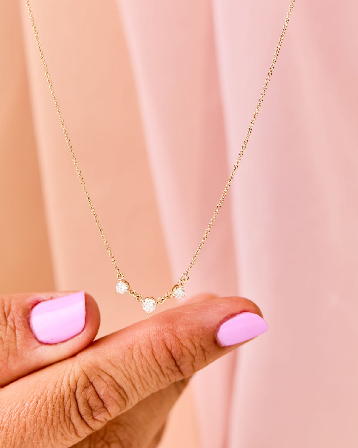 Fingers holding the Diamond Cascade Necklace against a pink curtain.