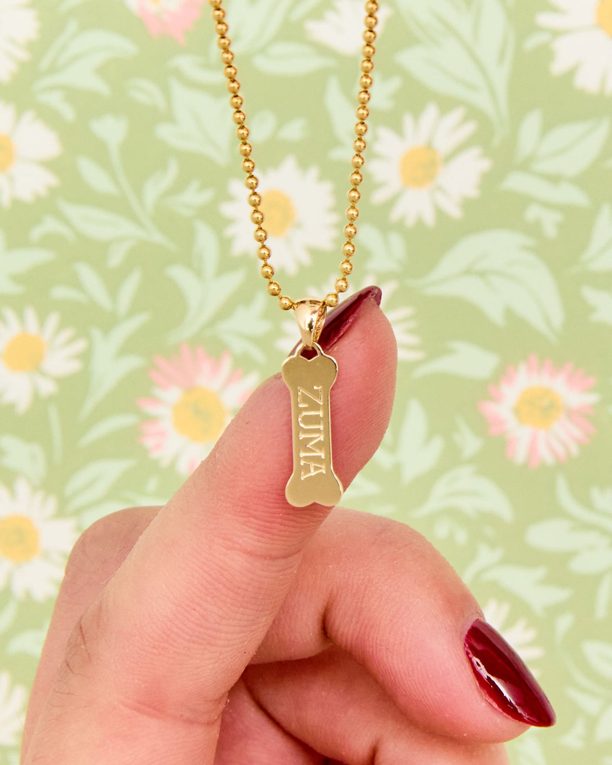 Fingertip holding the Dog Bone Signet Charm engraved ZUMA on a daisy print.